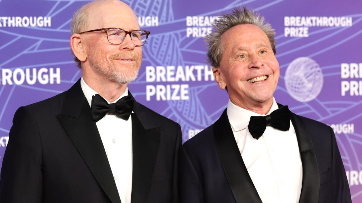 Two smiling men in black tuxedos pose on a purple Breakthrough Prize backdrop.