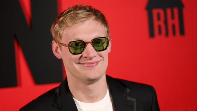 Portrait of a person with blond hair wearing green-tinted sunglasses and a black blazer, smiling against a red background with a black design.