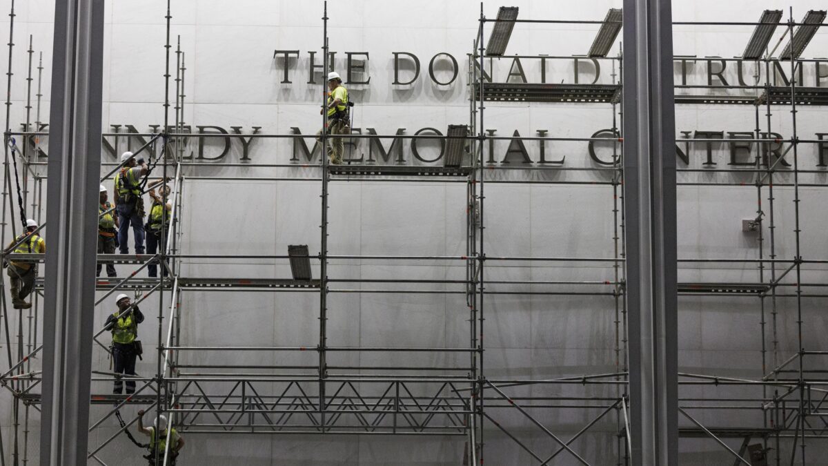 Construction workers in safety gear on scaffolding in front of a large white wall with partially visible lettering, urban construction site visible behind.