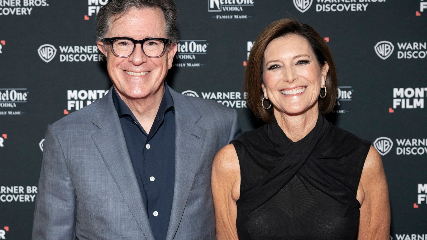 Two smiling adults posing together at a film event in front of a backdrop with logos; man in a gray suit and dark shirt, woman in a sleeveless black top.