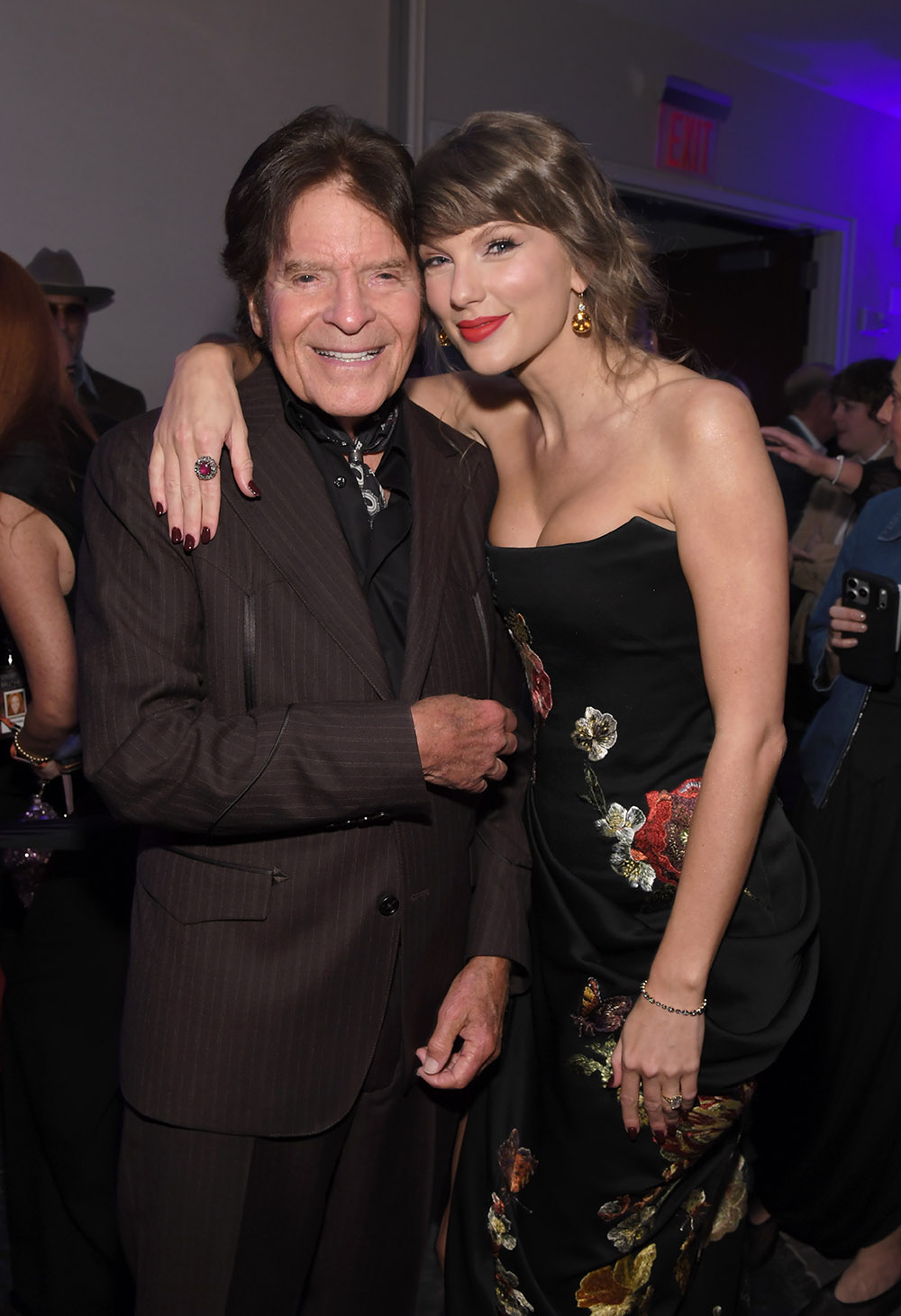 Smiling older man in a brown pinstripe suit with his arm around a young woman in a black floral gown at a party event.
