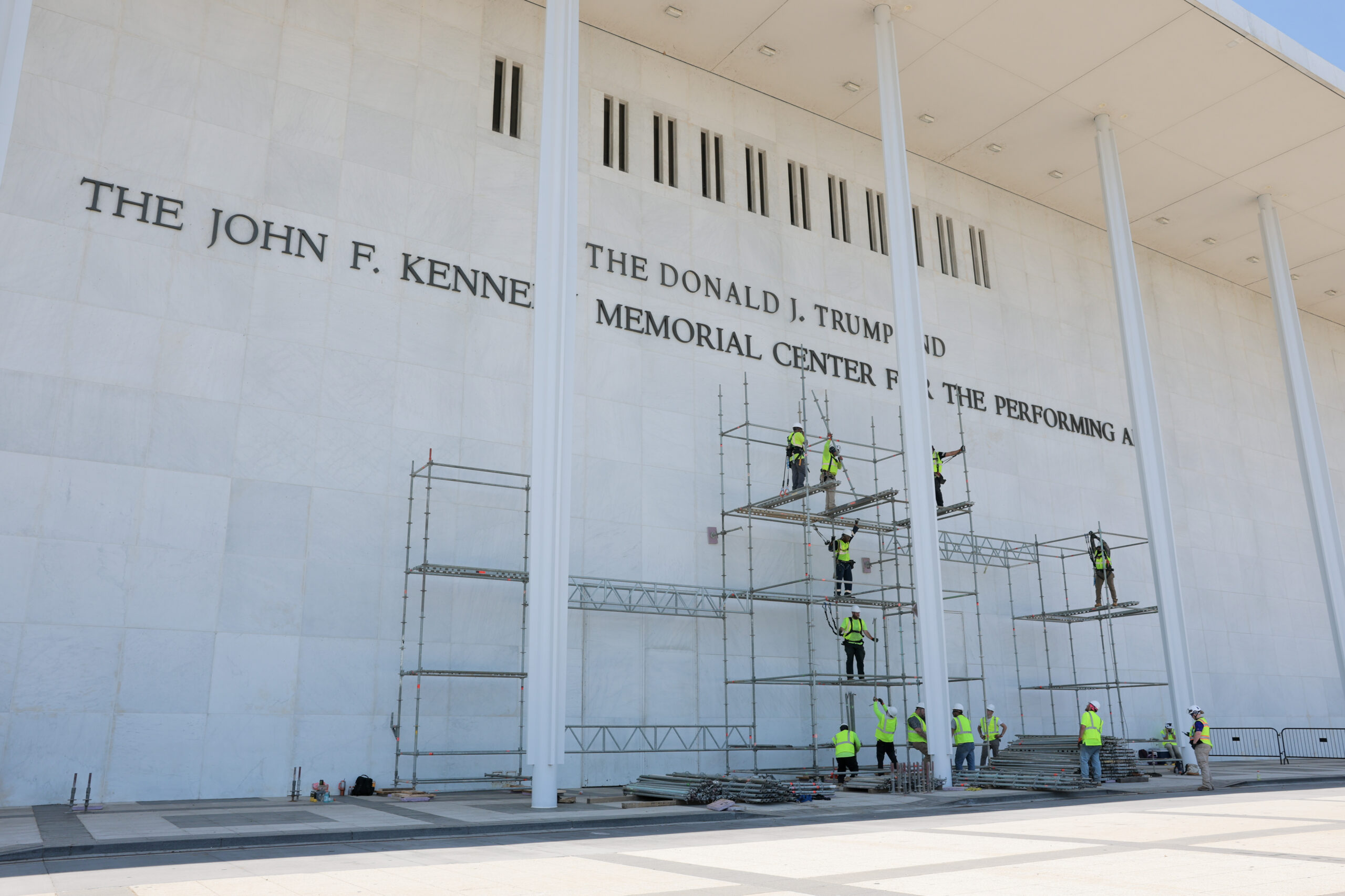 Construction workers on scaffolding outside the John F. Kennedy Center for the Performing Arts facade under a bright sky.