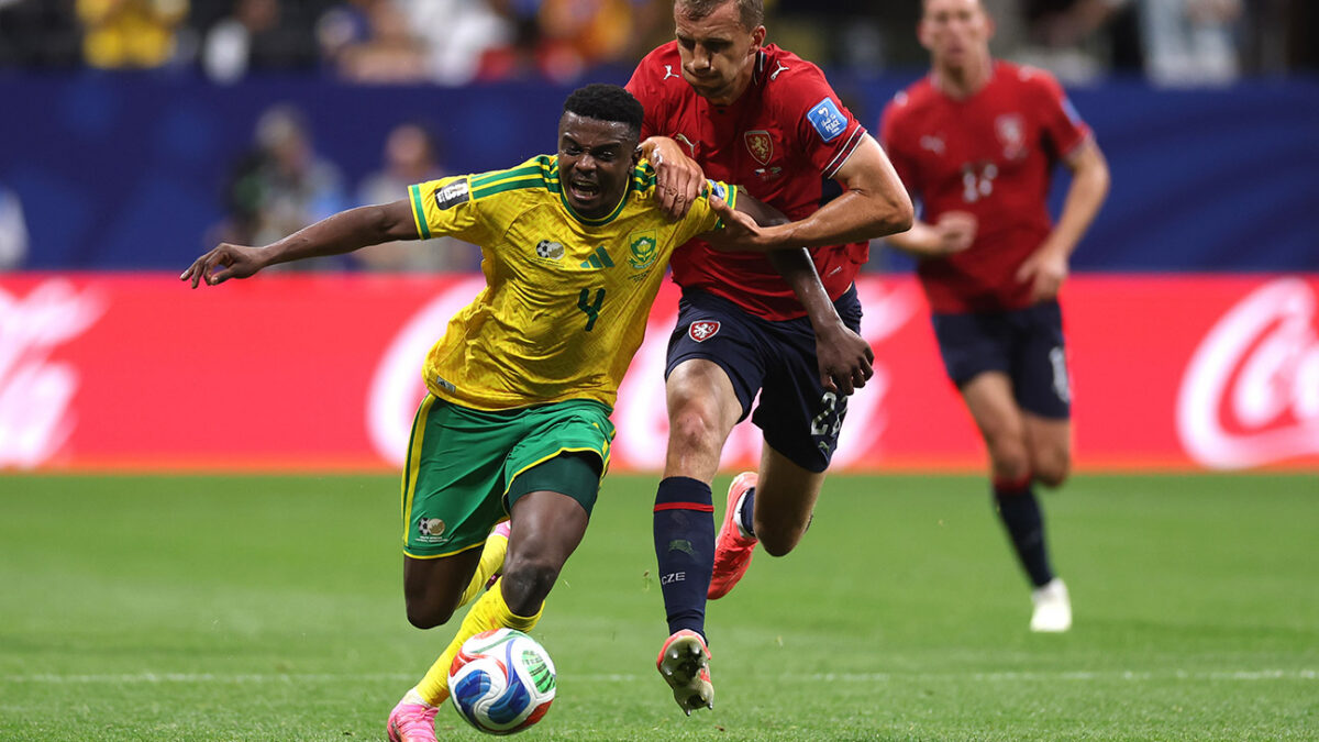 Two soccer players compete for the ball on a green pitch; one in yellow-green kit defends against a red-kit opponent.