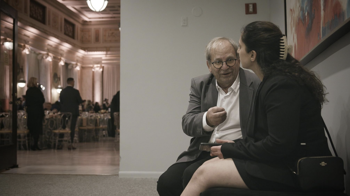 Older man in a suit and glasses speaks with a woman in a blazer as they sit by a wall at a formal event, banquet hall in the background.