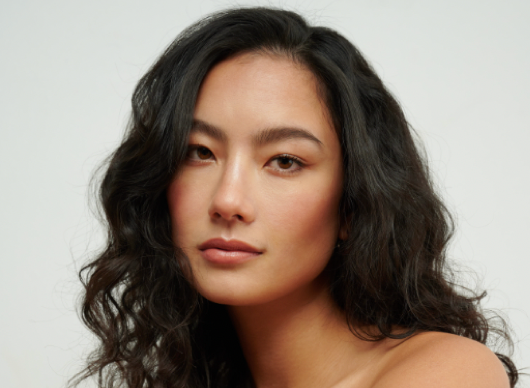 Close-up portrait of a young Asian woman with long, dark curly hair and a neutral expression against a light background, showcasing natural makeup.