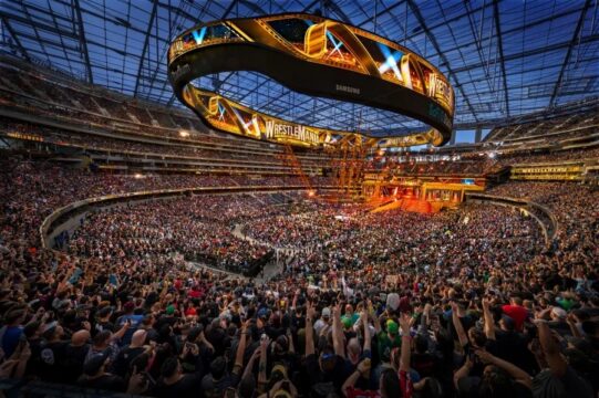 Large stadium filled with a cheering crowd watching a WrestleMania event with a glowing circular video board above the ring