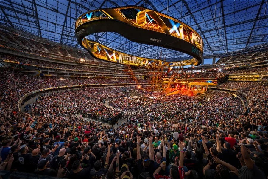 Large stadium filled with a cheering crowd watching a WrestleMania event with a glowing circular video board above the ring