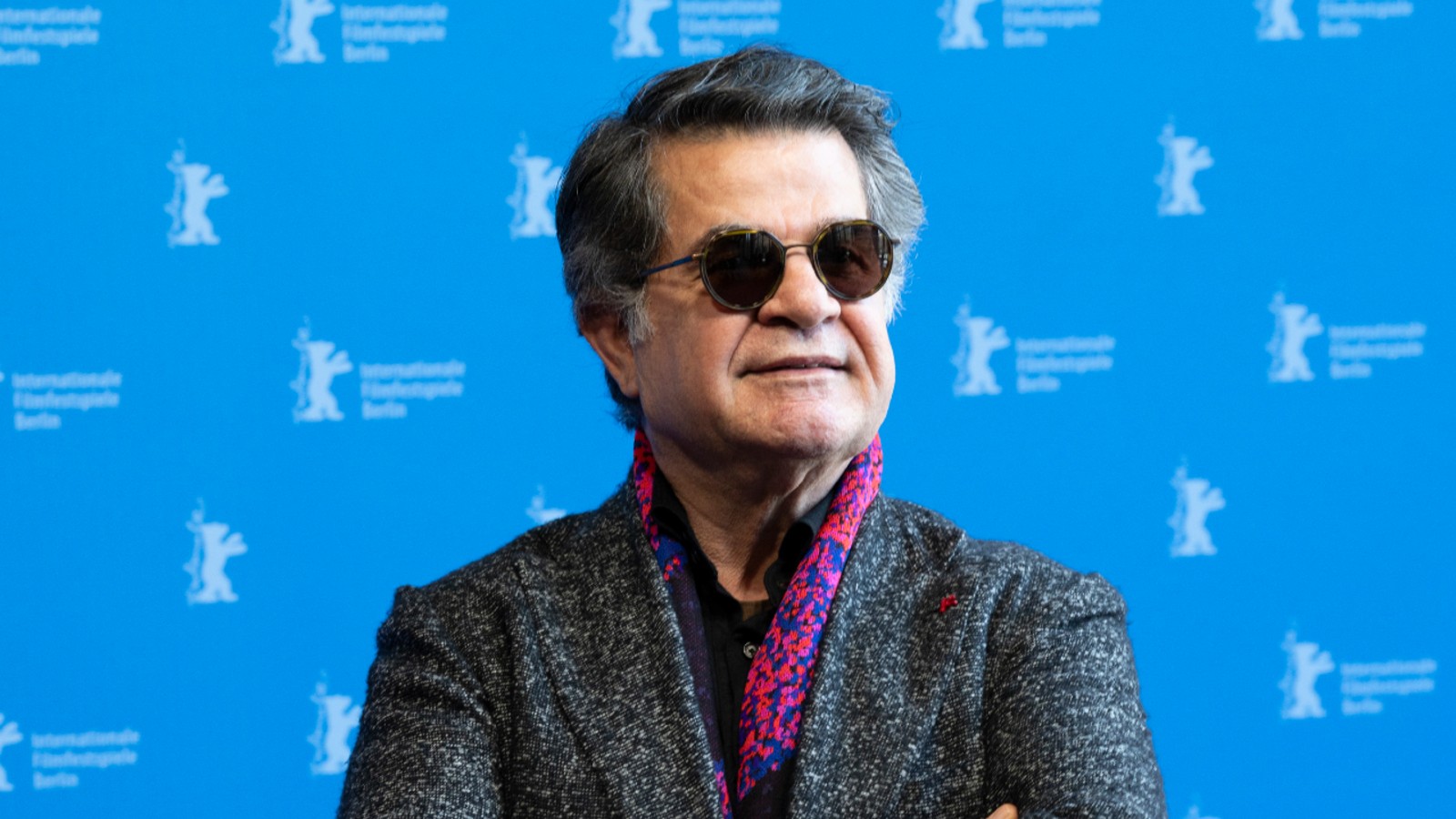 Man with gray hair and dark sunglasses in a textured blazer and purple scarf, posed in front of a blue Berlinale backdrop.