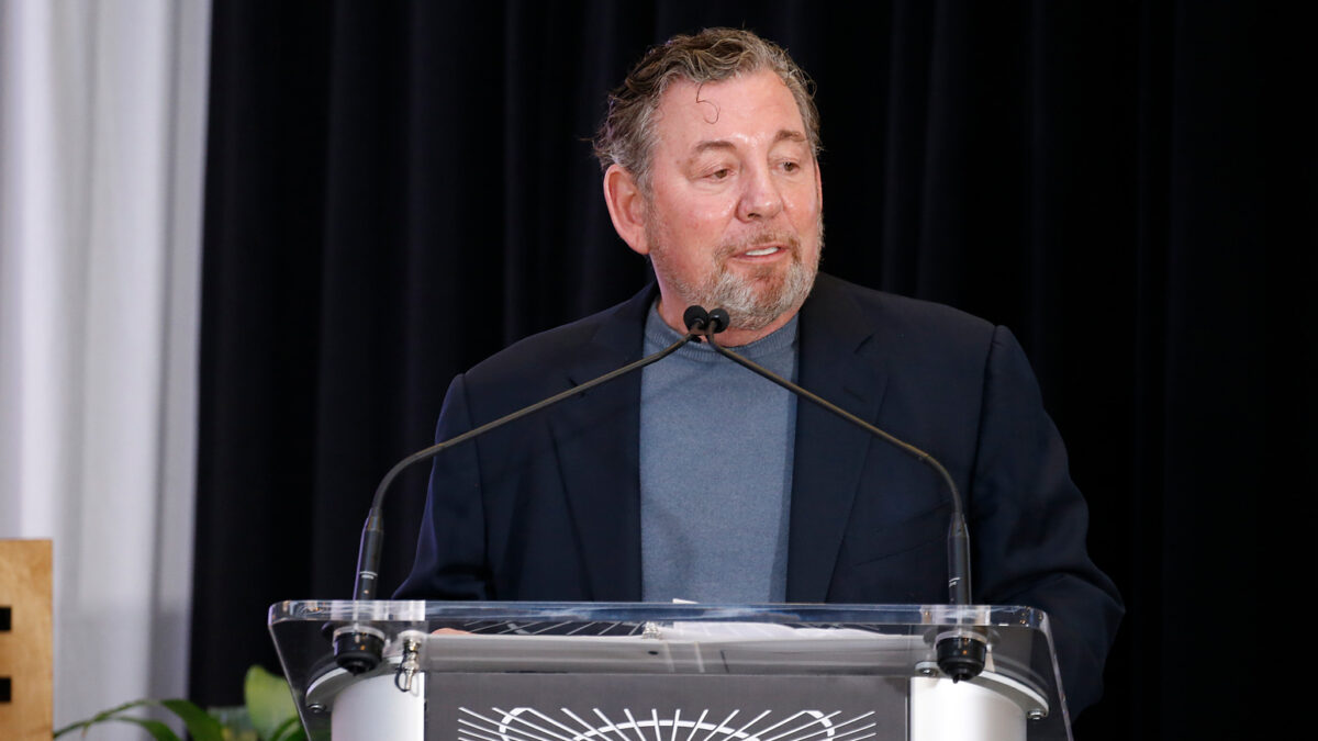 Man in a navy blazer speaks from a clear lectern with dual microphones on stage.