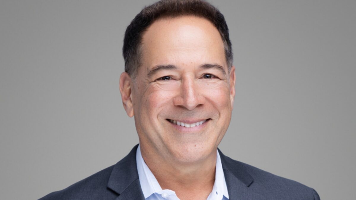 Head-and-shoulders portrait of a smiling man in a dark suit and light blue shirt against a gray backdrop.