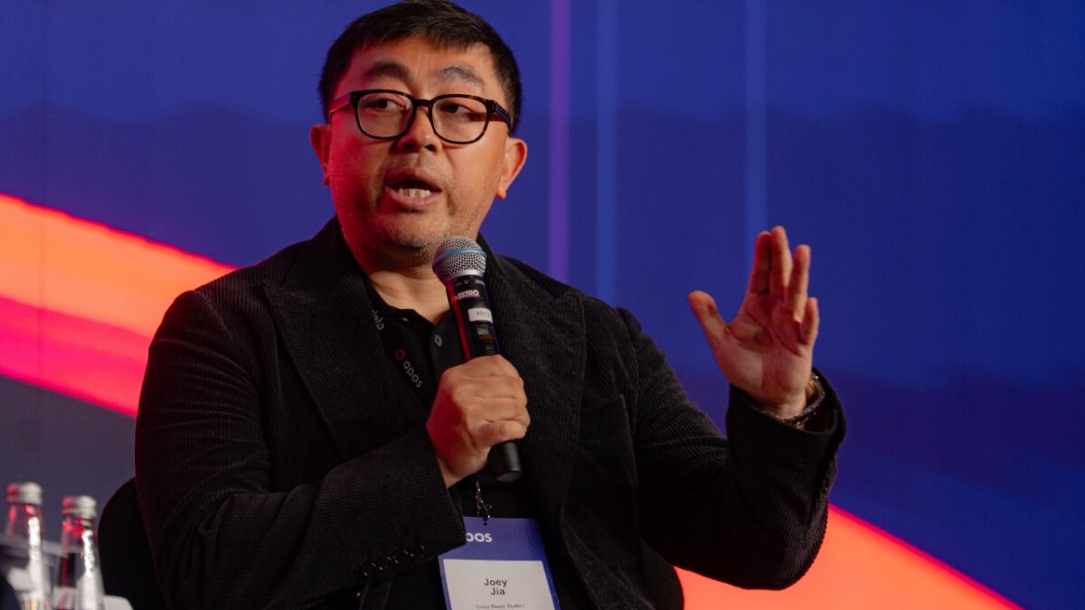 Man wearing glasses, speaking into a handheld microphone on stage, gesturing with his right hand
