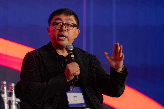 Man wearing glasses, speaking into a handheld microphone on stage, gesturing with his right hand