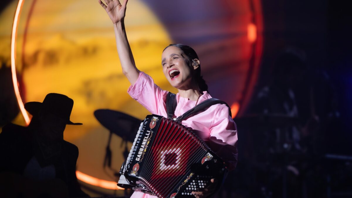 Female accordionist in a pink dress joyfully performs on stage, one arm raised to the crowd.