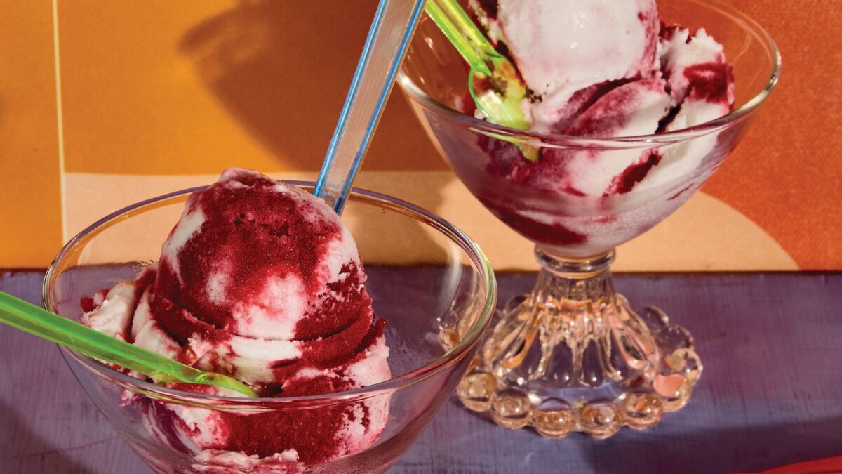 Two dessert glasses hold scoops of vanilla ice cream with berry sauce, colorful spoons, on a purple table with an orange tiled backdrop.”,
