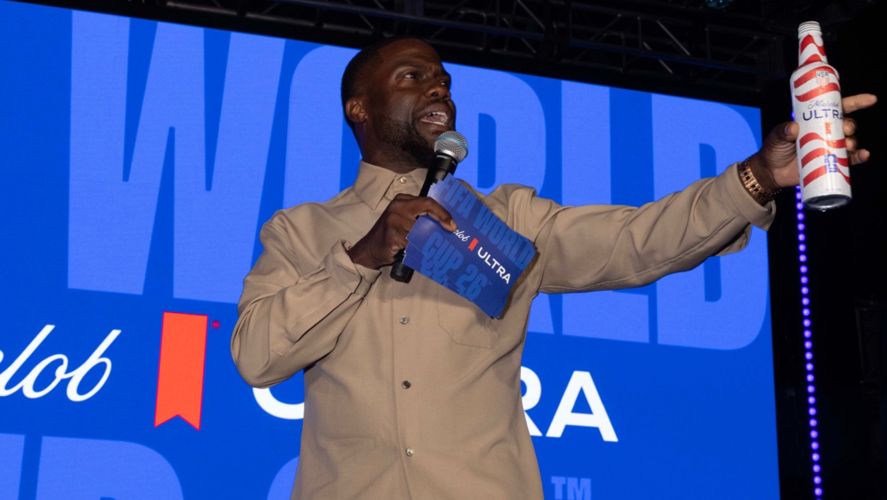 African American man on stage holding a microphone and a bottle of Ultra beer, with a blue backdrop behind him.