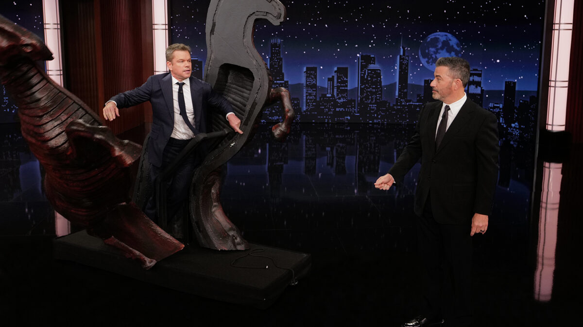 Two men in suits on a TV talk show set; one sits on a large red dragon-like sculpture while the other looks on, with a nighttime city skyline backdrop.