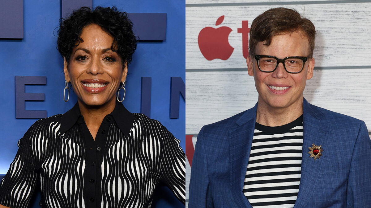 Two people posing: left is a smiling woman with short curly hair in a black-and-white patterned blouse; right is a smiling person in a blue blazer, striped shirt, glasses, wearing a sun-shaped brooch with a red heart.
