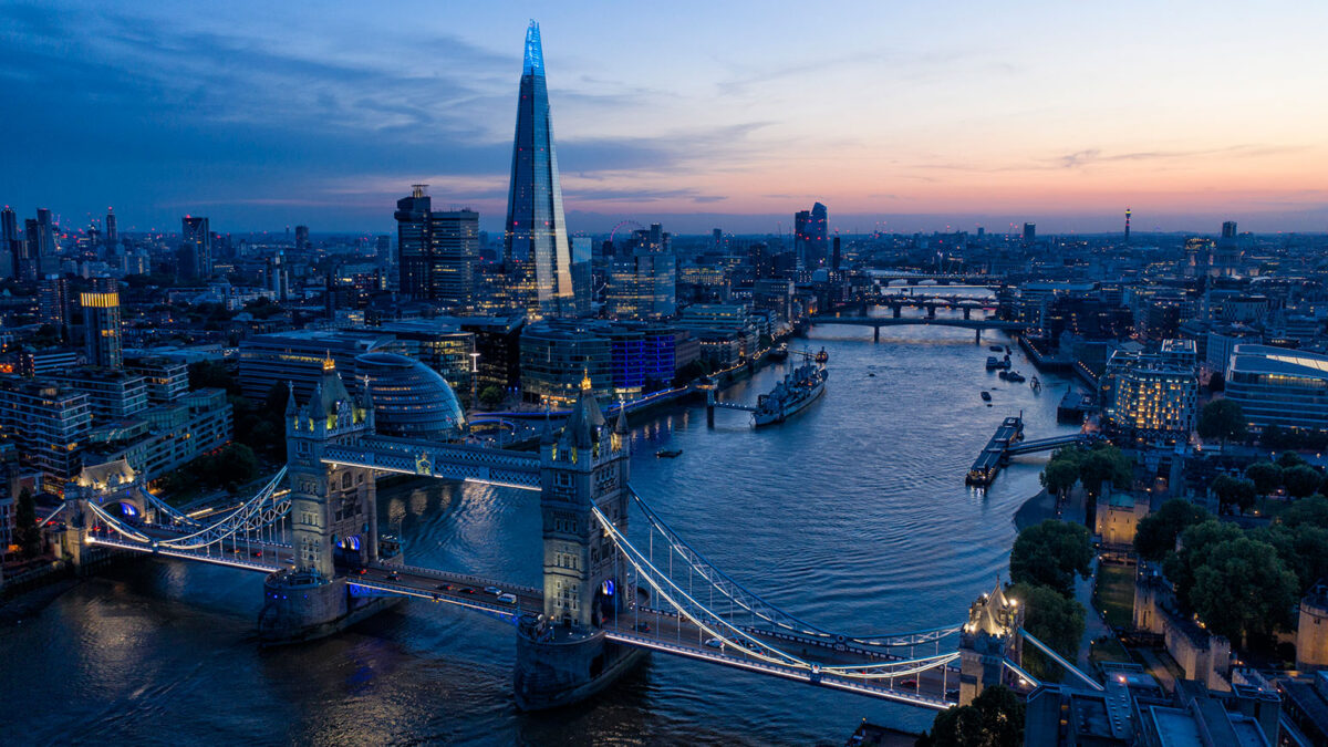 Aerial view of Tower Bridge spanning the River Thames at dusk, with the London skyline including The Shard in the distance.