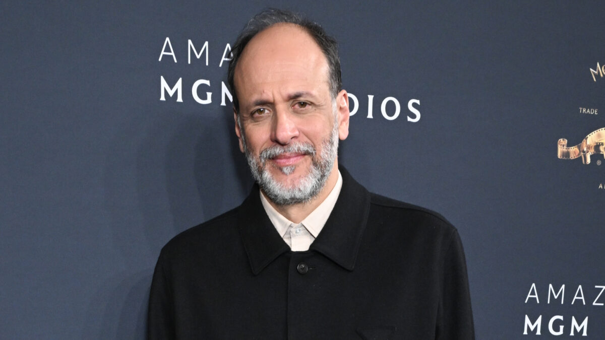 Smiling man with a gray beard wearing a black coat at a blue MGM backdrop.