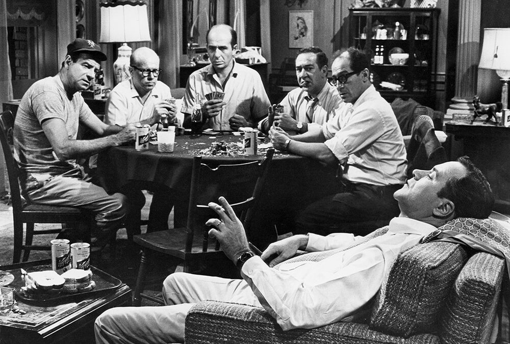 Five men around a cluttered table playing cards; one man leans back in a chair watching, in a living room setting with drinks and snacks nearby.