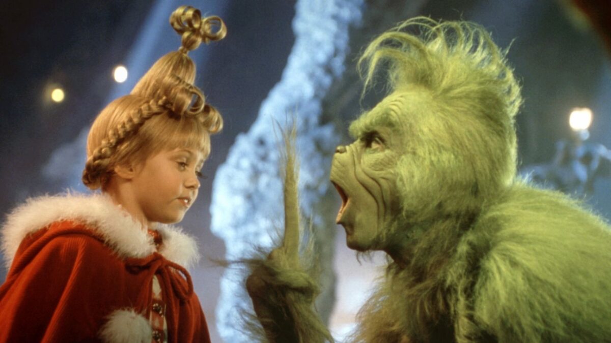 A young girl in a red fur-trimmed cloak talks with a green, furry creature in a fantasy scene with a snowy background.