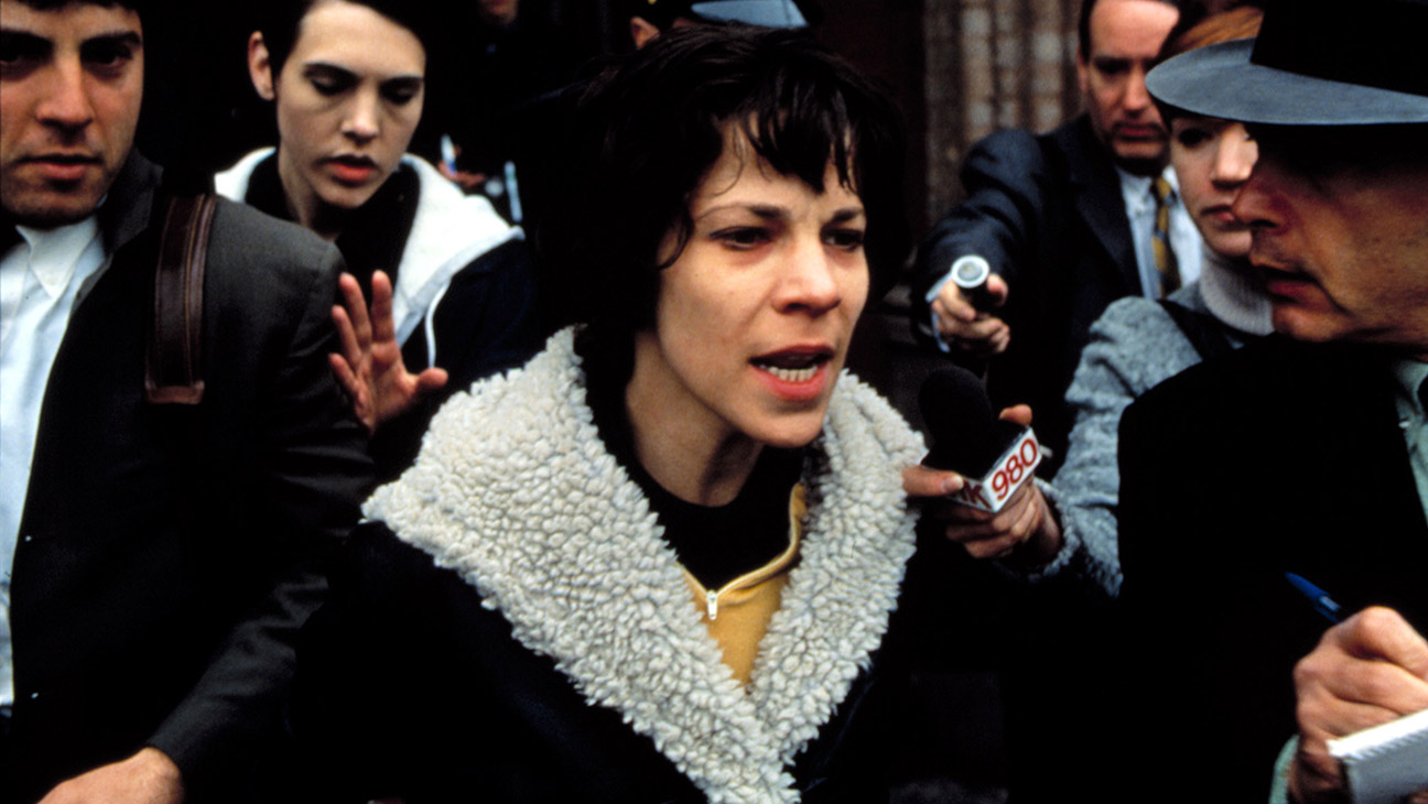Woman with short dark hair in a cream shearling coat speaks into a microphone as reporters crowd around her.