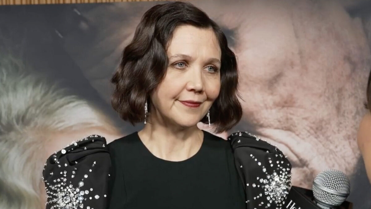 Smiling woman with a short dark bob in a black beaded dress speaks into a microphone at an event, backdrop blurred behind her.
