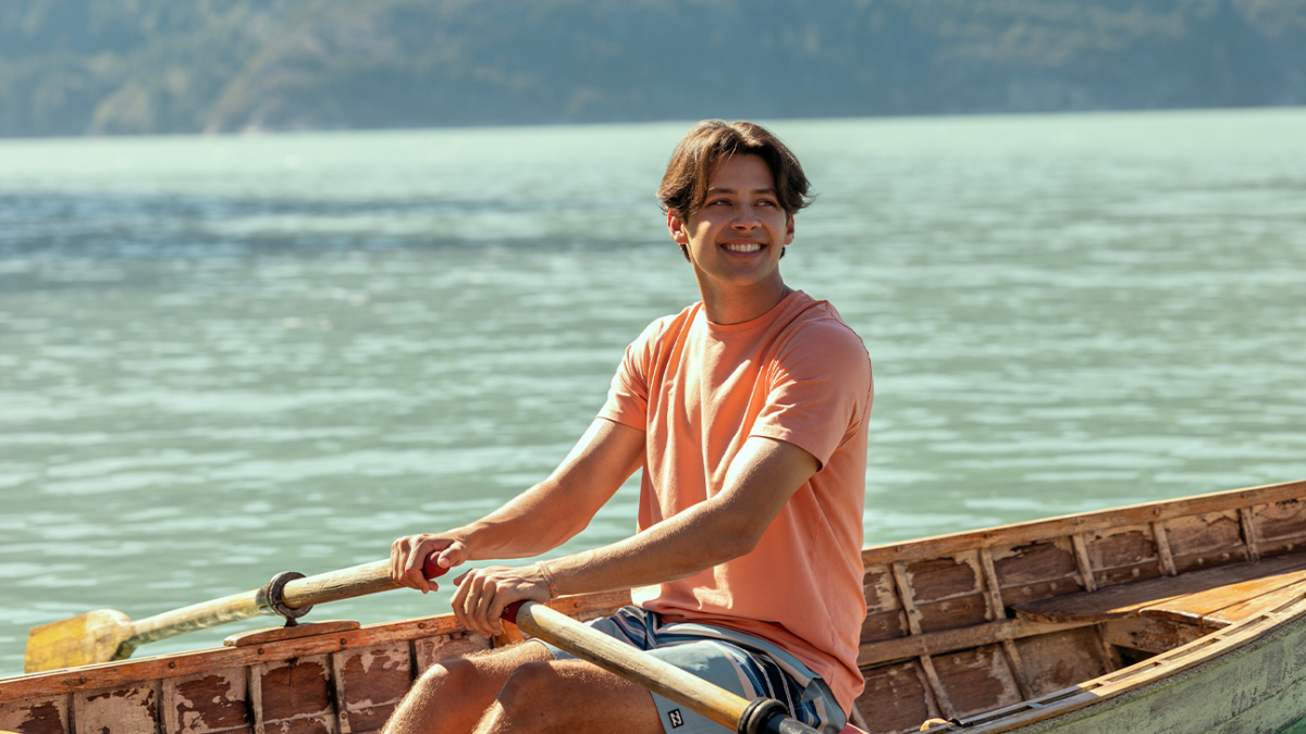Young man in a peach T-shirt smiles while rowing a weathered wooden boat on a calm, greenish lake with hills in the distance.