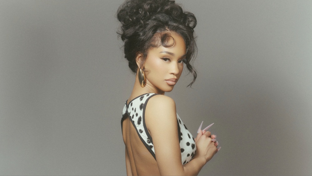 Portrait of a woman with dark curly hair wearing a backless white dress with black spots, looking over her shoulder at the camera, against a gray backdrop, wearing gold hoop earrings.