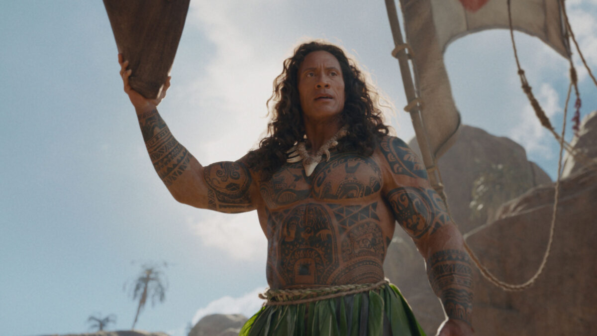 Man with extensive Polynesian tattoos raises a padded rope while standing on a ship rigging against a rocky tropical backdrop.