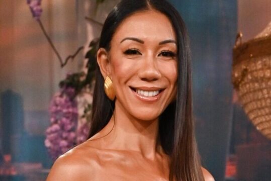 Smiling Asian woman with long dark hair and gold earrings, posing in a studio setting.