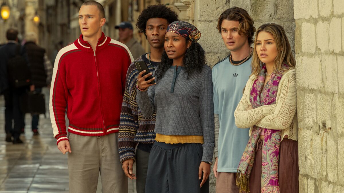 Five young adults stand closely together on a city street, leaning against a stone wall and looking off-camera, with one person holding a smartphone.