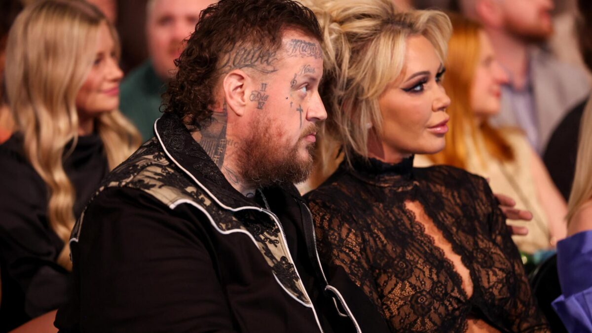 Man with many facial tattoos and beard seated next to a woman in a black lace outfit at an event, audience in background