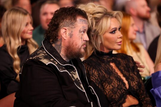 Man with many facial tattoos and beard seated next to a woman in a black lace outfit at an event, audience in background
