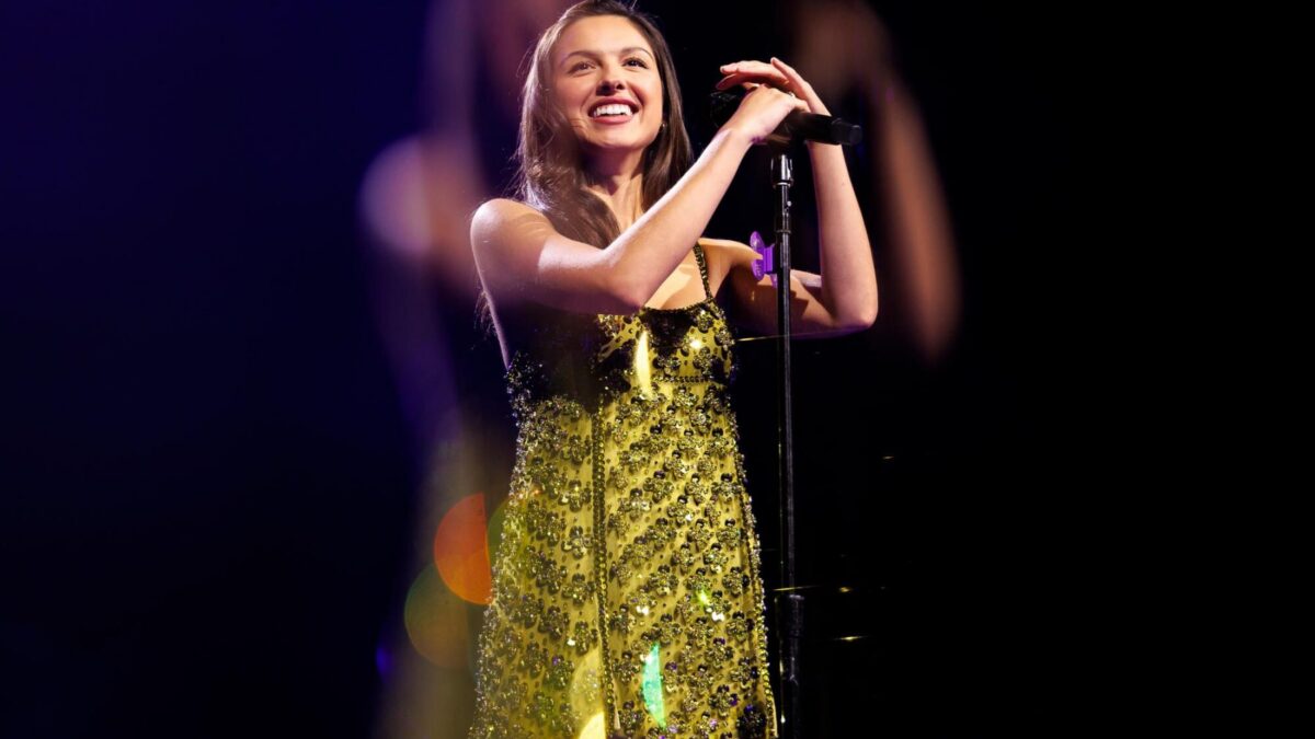Female singer smiling while singing into a microphone on stage, wearing a sparkling yellow beaded dress under stage lights