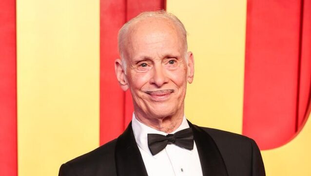 Elderly man in a black tuxedo and bow tie, smiling at the camera, against a red and yellow backdrop at a formal event.