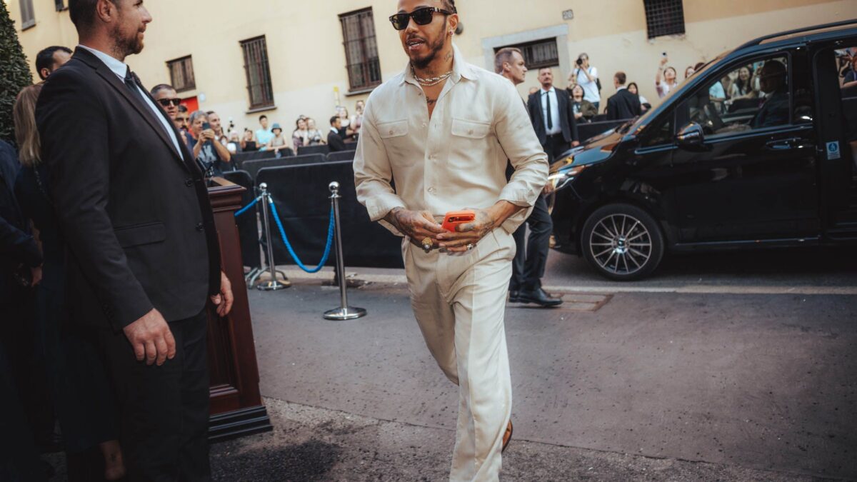 Man in a beige jumpsuit and sunglasses walks toward cameras at a red-carpet event, with fans and security barriers in the background and a black van nearby.