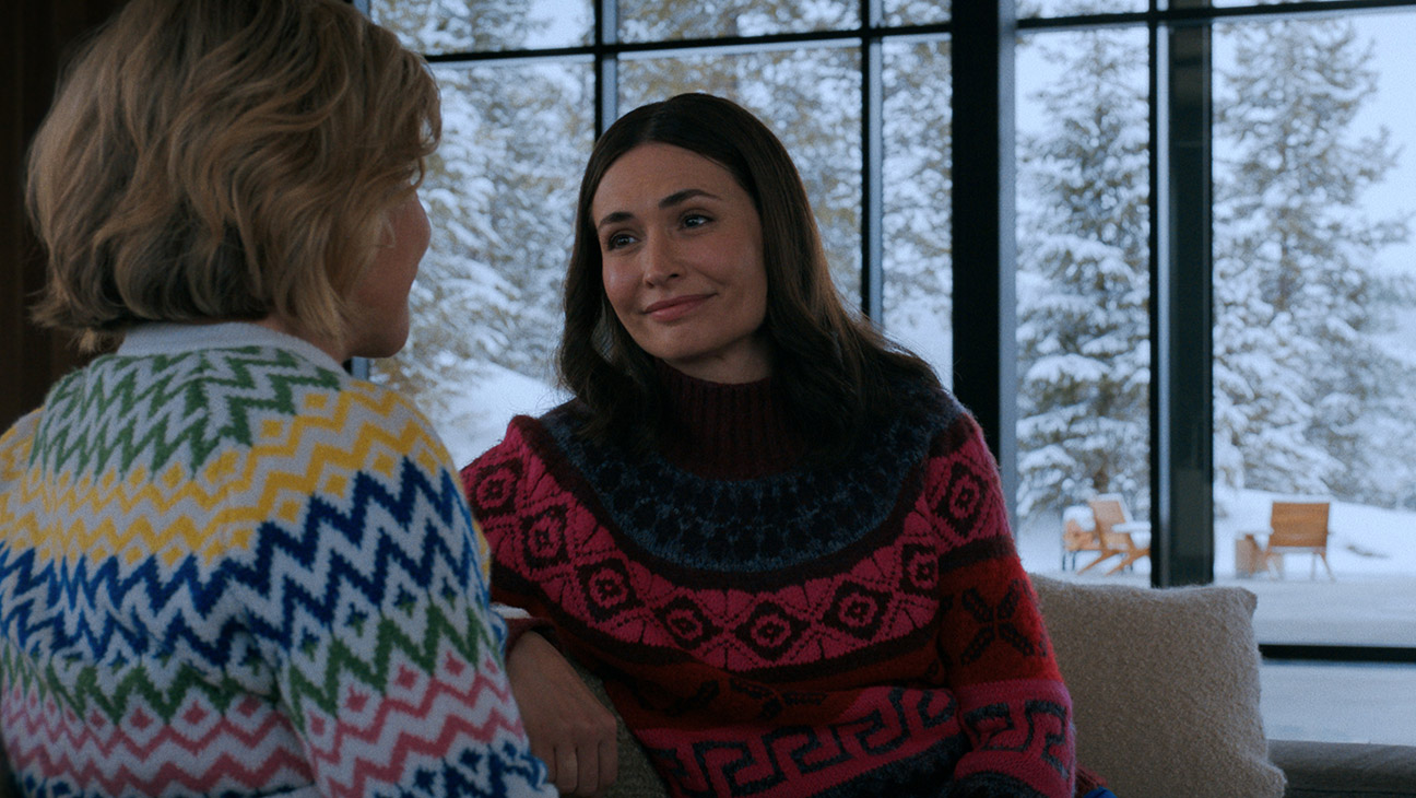 Two women sit and chat indoors by a large window; one wears a multicolored zigzag sweater, the other in a pink and blue patterned sweater, smiling at each other in a cozy setting with a snowy scene outside.