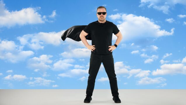 Man in black shirt and sunglasses stands with hands on hips, cape billowing behind him against a blue sky with clouds.