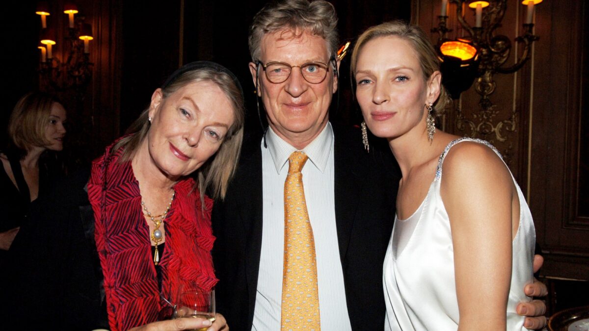 Three adults posing at a formal event: a woman in a red blouse, a man in a suit with an orange tie, and a woman in a white satin dress; chandeliers glow in the background.