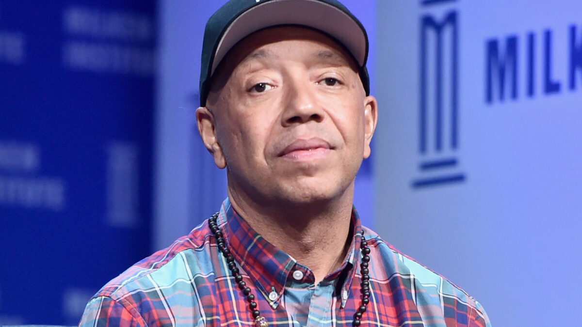 Man wearing a dark baseball cap, plaid shirt, and beaded necklace, with a lavalier mic clipped to his shirt on a stage backdrop.