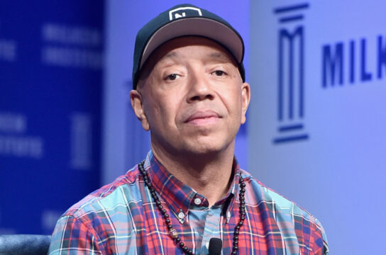 Man wearing a dark baseball cap, plaid shirt, and beaded necklace, with a lavalier mic clipped to his shirt on a stage backdrop.