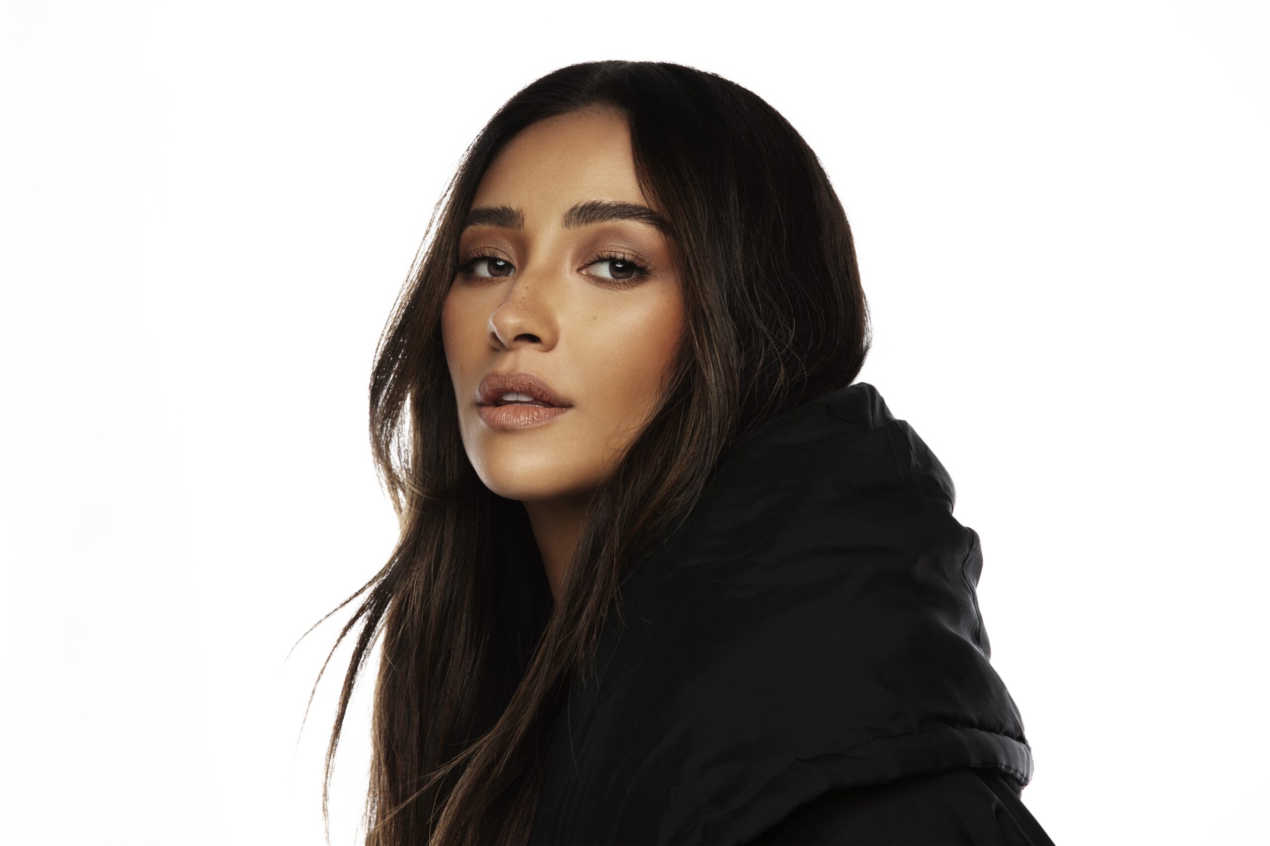 Close-up portrait of a woman with long dark hair, wearing a black jacket, looking slightly over her shoulder against a white background.