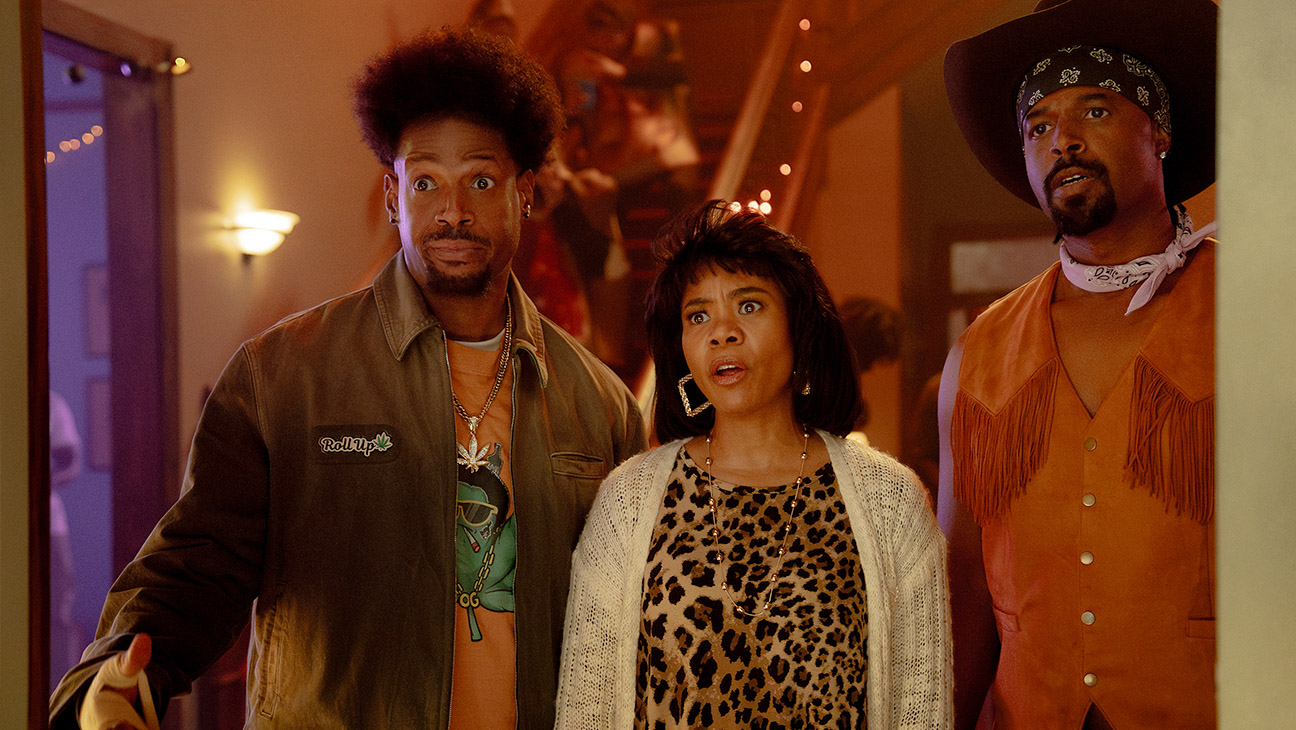 Three partygoers look surprised in a warmly lit room: left man in olive jacket, center woman in leopard print, right man in orange fringe vest.