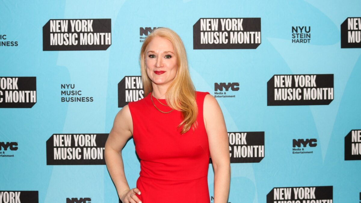 Woman in a red sleeveless dress posing in front of a blue 'New York Music Month' backdrop with logos.
