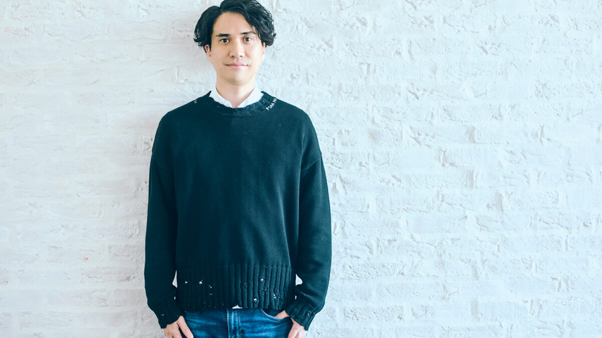 Young man with dark hair wearing a black sweater and jeans, standing against a white brick wall.