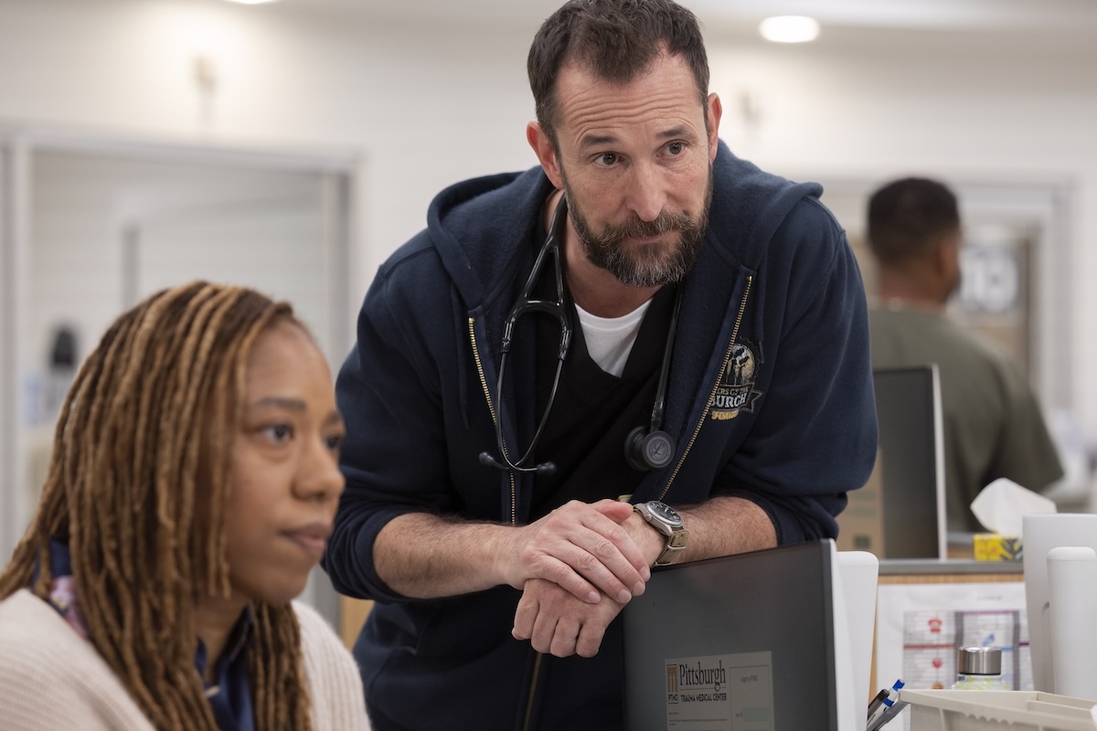 A doctor with a stethoscope leaning over a desk, talking to a patient in a clinic.