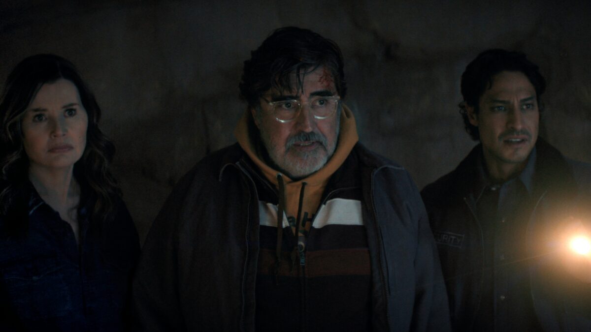 Three adults stand closely in a dark cave, illuminated by a flashlight in the right foreground, looking serious or concerned.