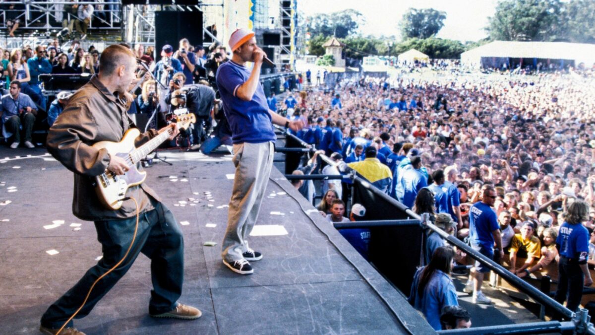Band performs on a large outdoor stage while a dense audience crowds the front barrier outside a festival venue.