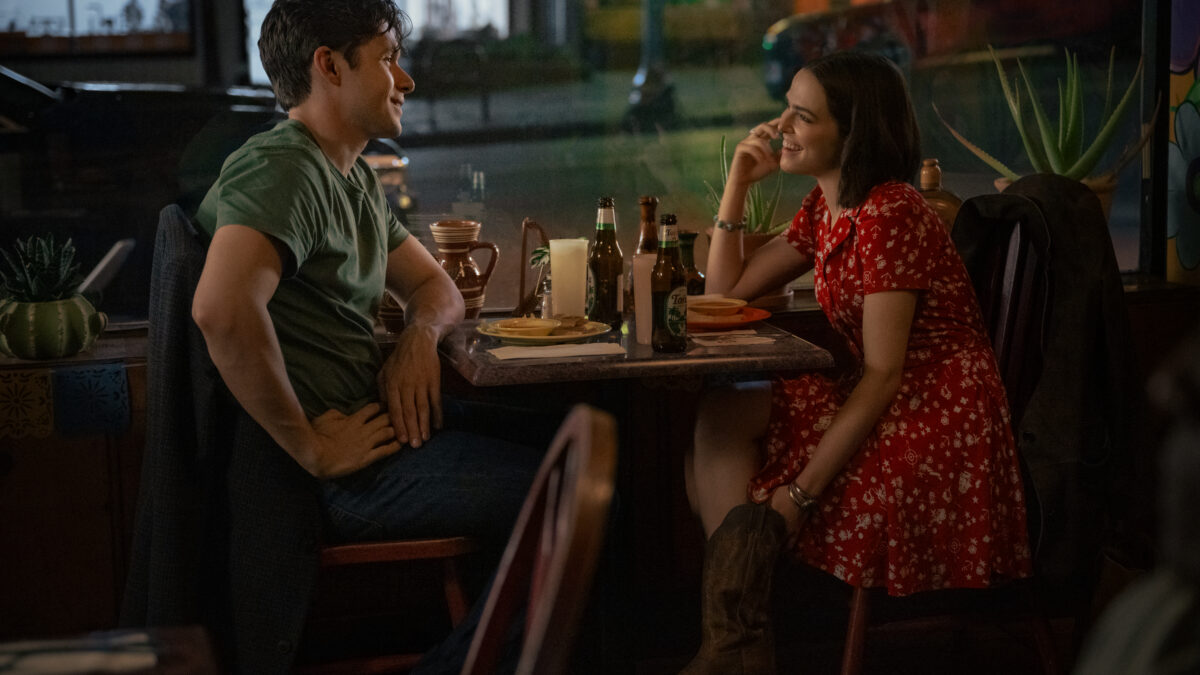 Man in a green shirt and a woman in a red dress sit across from each other at a dimly lit restaurant table, smiling and talking.